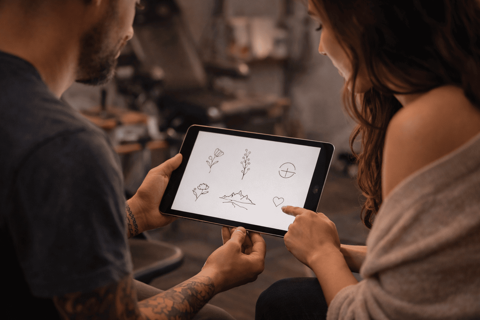 Tattoo-Künstler und Kundin sitzen gemeinsam im Tattoo-Studio und betrachten auf einem Tablet verschiedene Fineline-Tattoo-Motive, darunter florale, geometrische und minimalistische Linienzeichnungen, in ruhiger, warmer Studio-Atmosphäre.