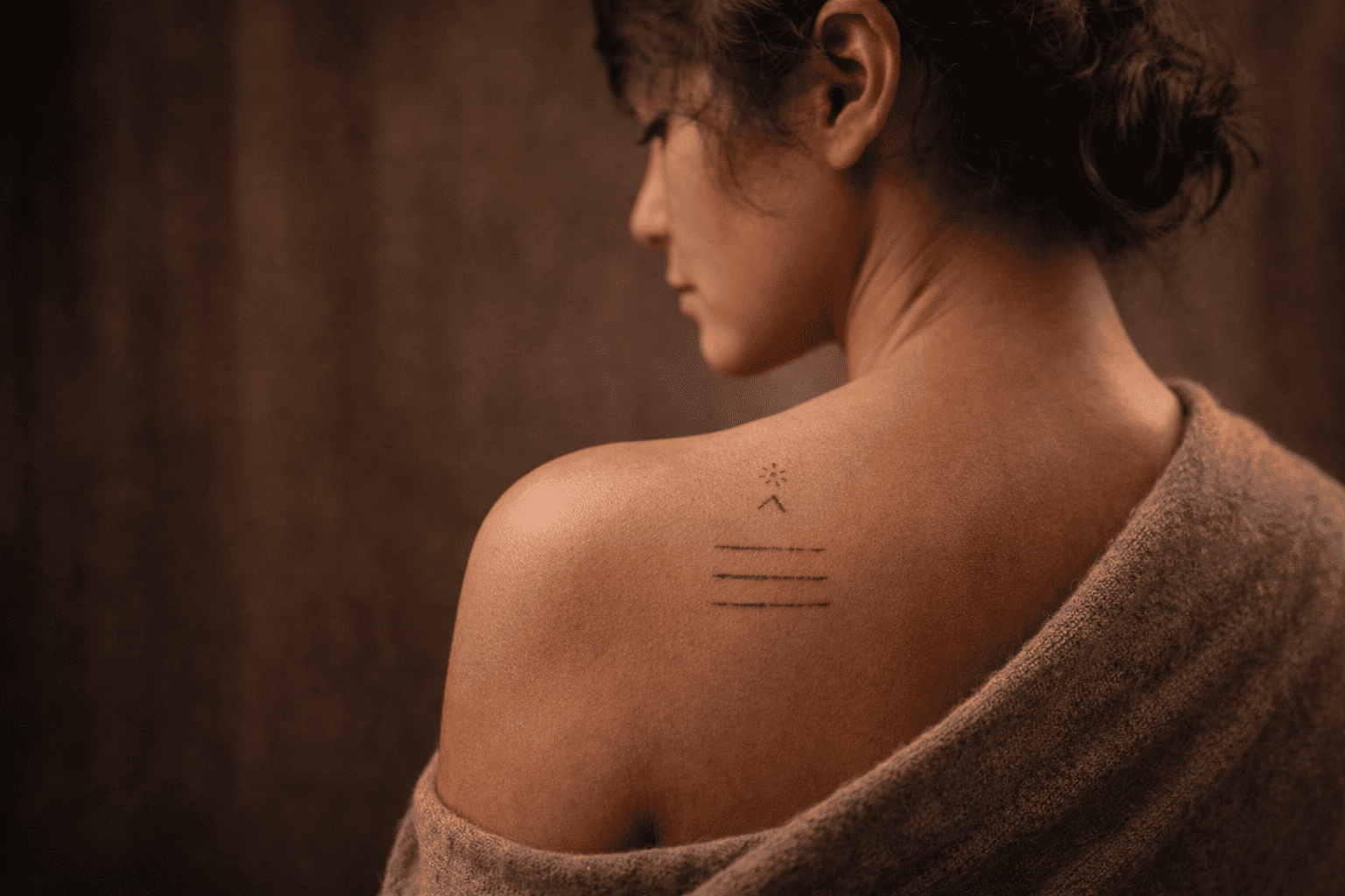 Ruhige Portraitaufnahme einer Person mit entspannter Körperhaltung, auf deren Haut ein dezentes Fineline-Tattoo mit feinen Linien sichtbar ist, dargestellt in warmer, natürlicher Lichtstimmung mit Fokus auf Mensch und Handwerk.