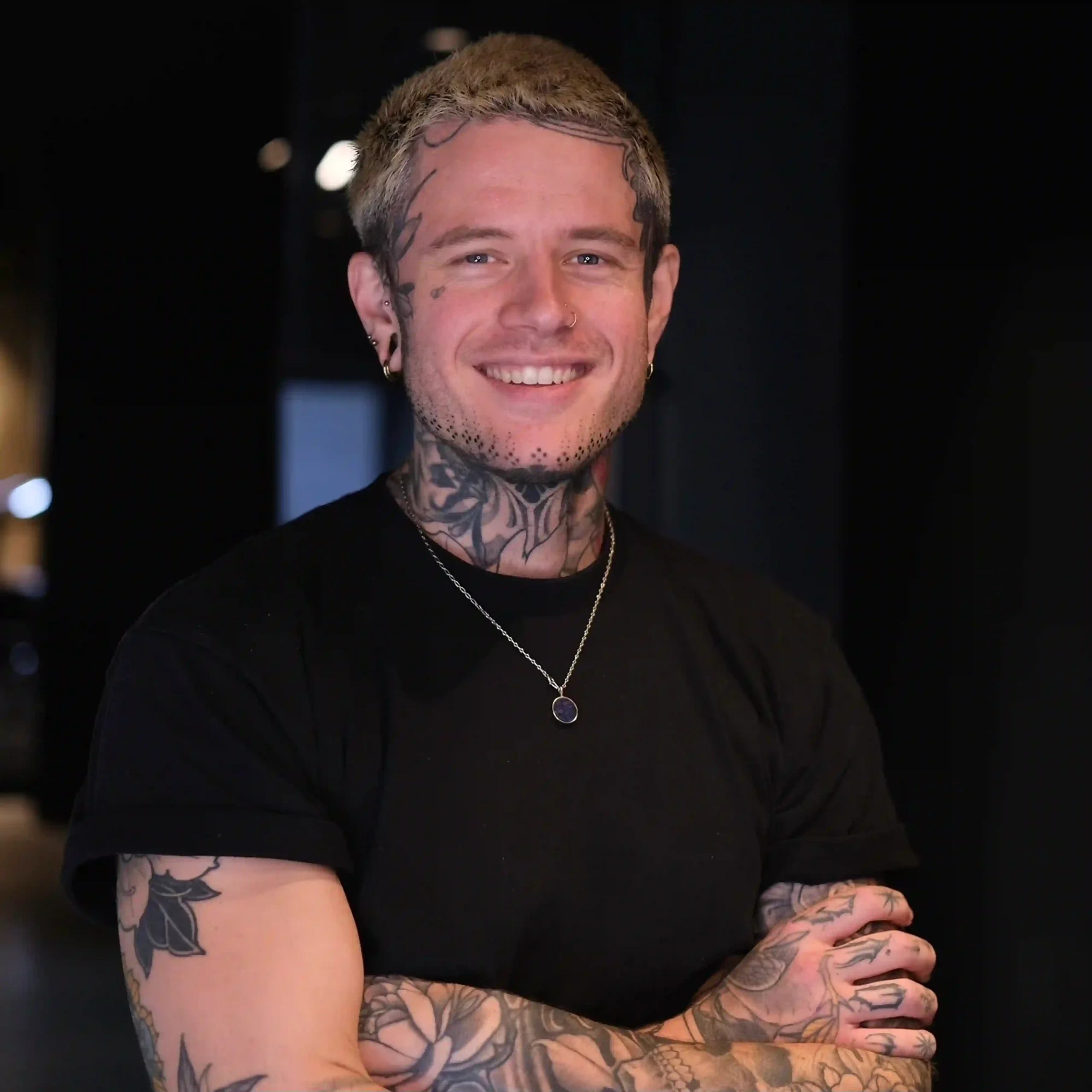 Lächelnder, tätowierter Mann in schwarzem T-Shirt im modernen Hood7-Tattoo-Studio, mit Hals- und Arm-Tattoos, verschränkten Armen und freundlicher Ausstrahlung.