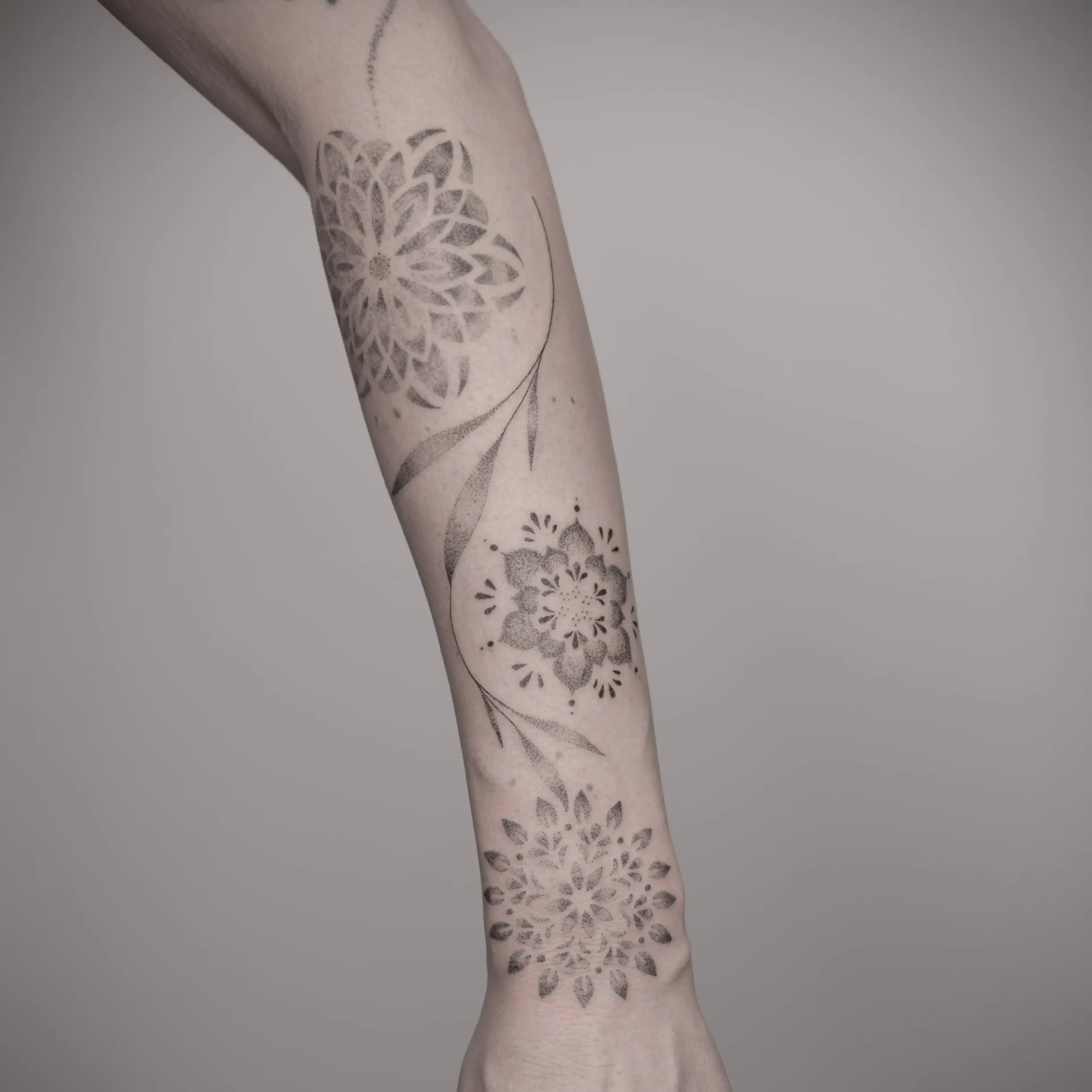 Feines Fineline-Tattoo auf dem Unterarm mit zarten Blumen und Ornamenten in schwarzgrauer Schattierung.